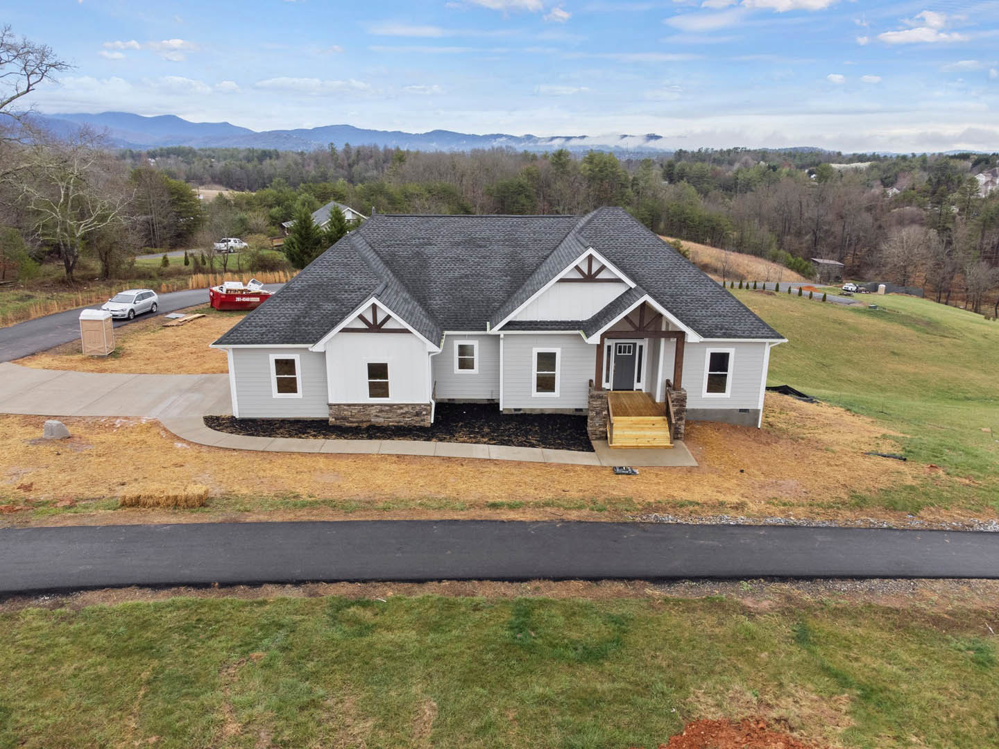 Two-story home with grey roof, white siding, wooden porch deck, concrete driveway, green grass lawn, trees in background, parked car and red boat near roadside