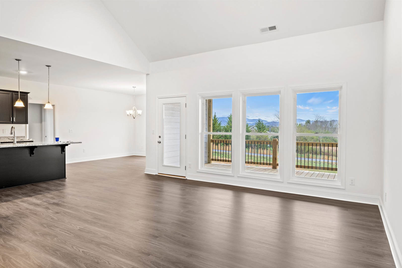 Spacious empty room featuring hardwood floors, black accent wall with white countertop, white door with silver knobs, large window overlooking trees, deck access, and wooden