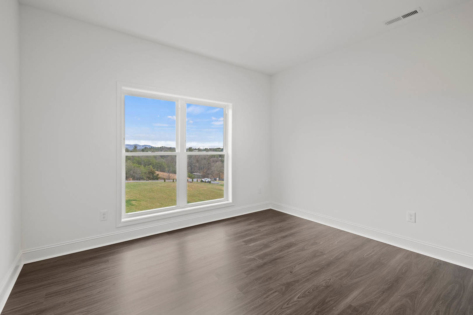 Sunlit room featuring wide plank hardwood flooring, white plaster walls, and a large window overlooking a grassy field and distant trees.
