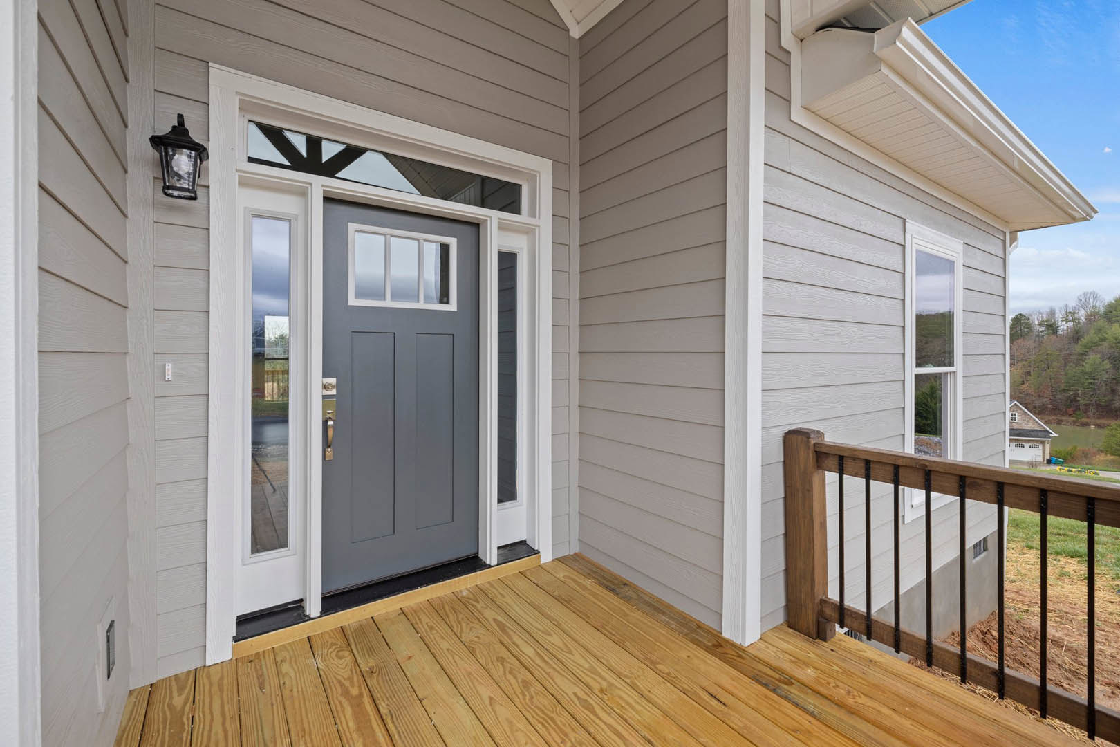 Grey door with glass panes set in white siding, wooden deck with railing, outdoor lamp mounted beside entry, open door revealing wood flooring inside.