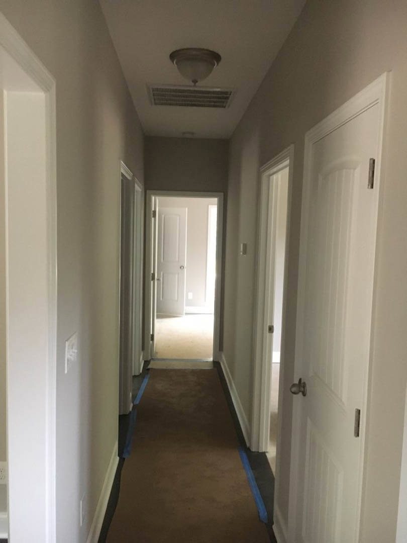 Carpeted hallway with white walls, ceiling light fixture, blue painter’s tape along baseboards, white door, and wall vent