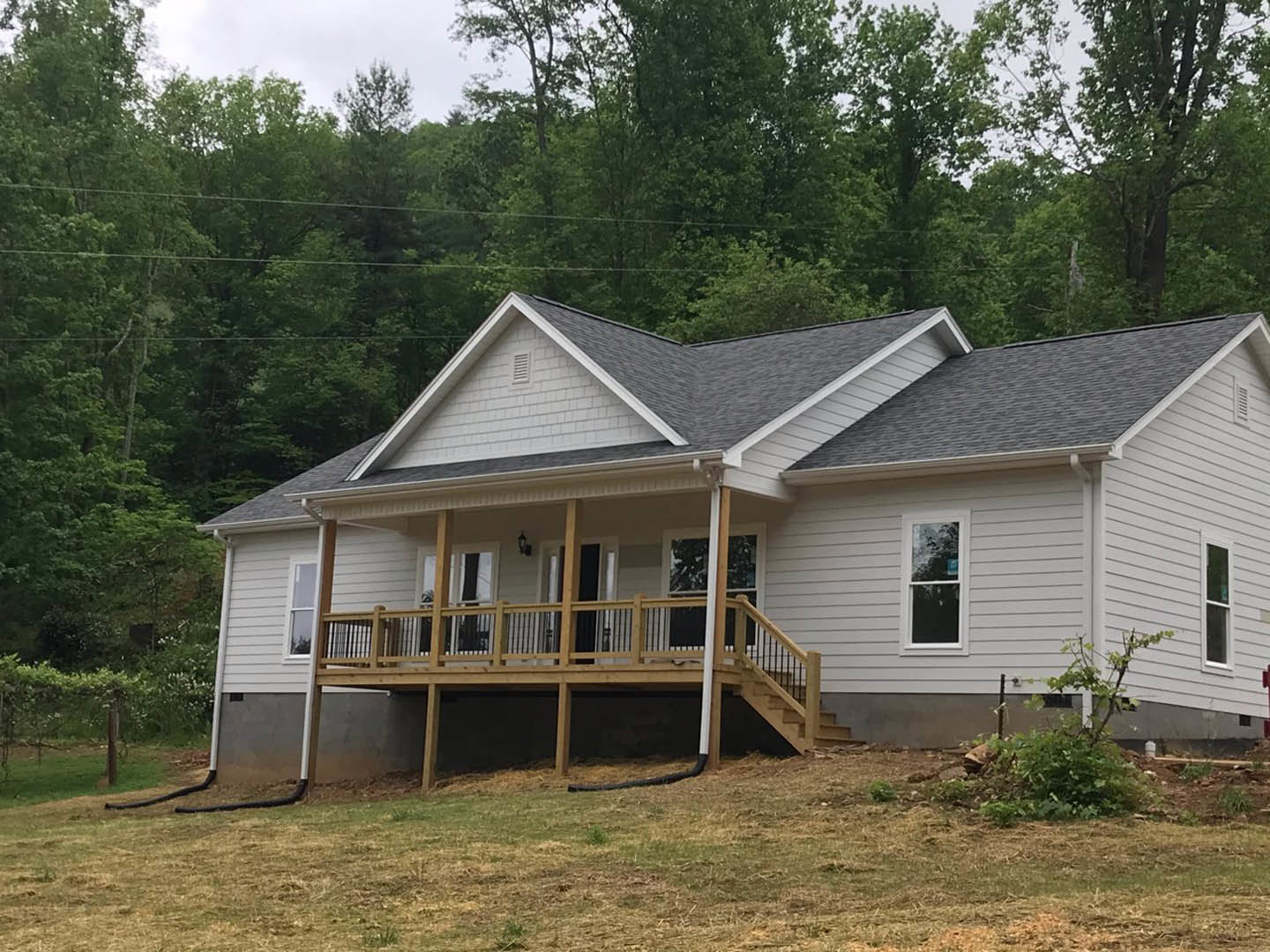 Two-story house with light siding, covered porch, wooden deck with railing, stairs leading to yard, large windows, surrounding trees, black pipe on ground, power lines in