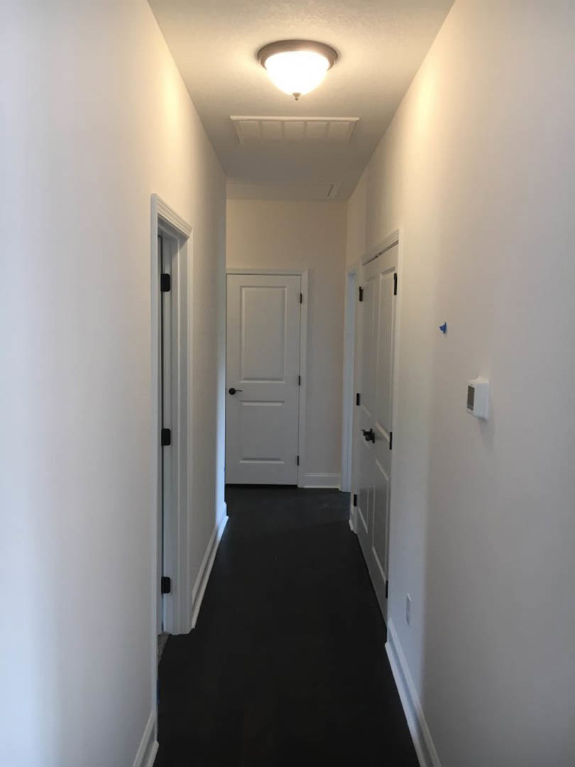Hallway with white plaster walls, multiple white doors featuring black handles, dark carpet flooring, and a ceiling-mounted light fixture
