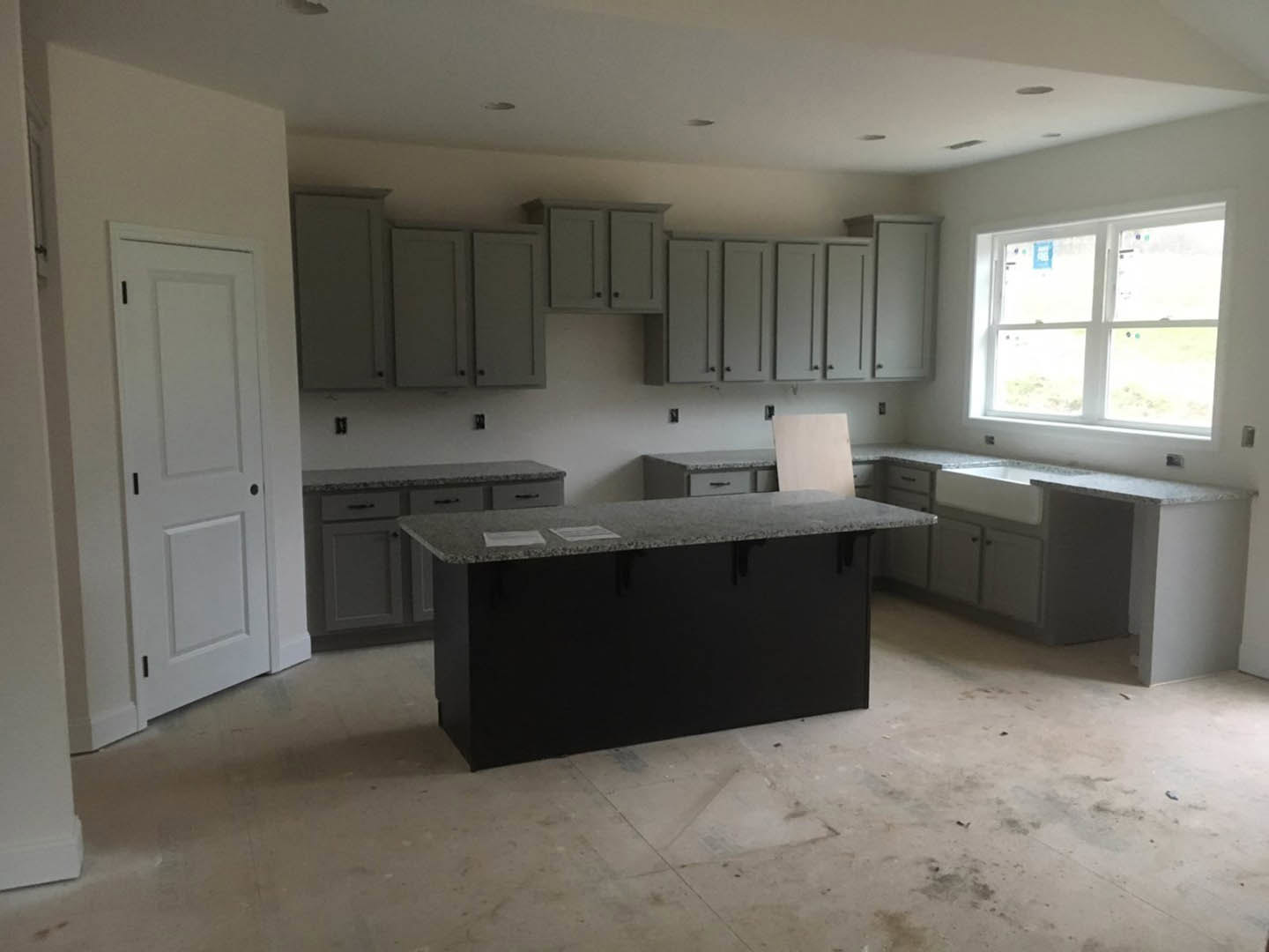 Grey shaker cabinets with black hardware, white tile backsplash, quartz countertop scattered with papers, stainless steel sink beneath a white-framed window, white doors with black