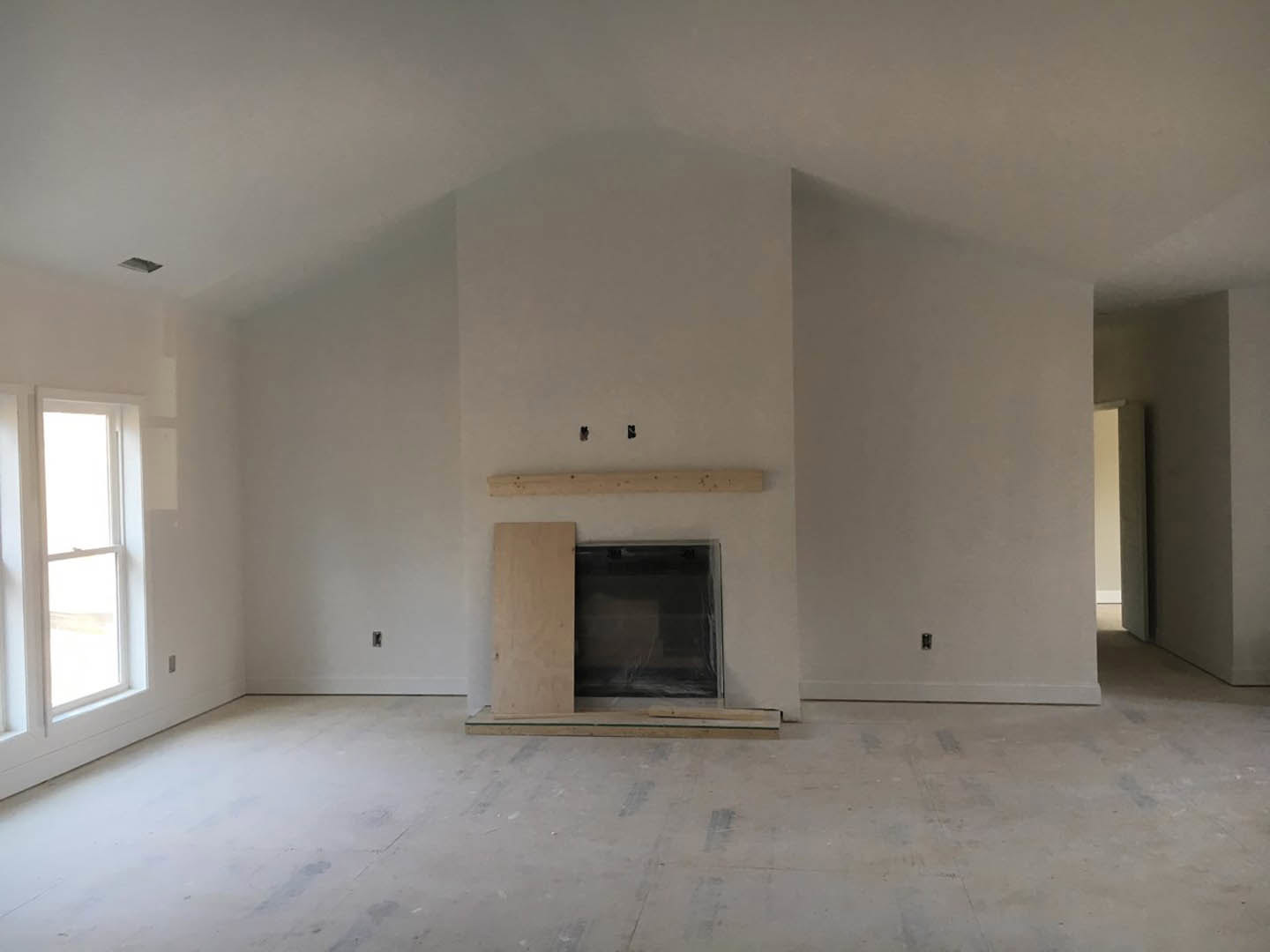 Living room with white-framed window covered in plastic wrap, built-in wood shelf, plaster walls, and fireplace; unfinished flooring with exposed hole near wall.
