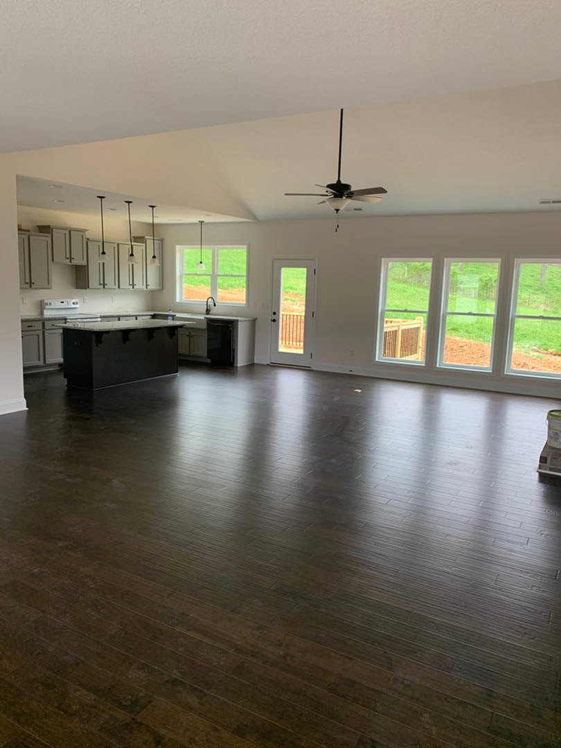 Spacious open floor plan featuring wood laminate flooring, modern kitchen with black cabinetry and white countertops, ceiling fan with light fixture, large windows overlooking
