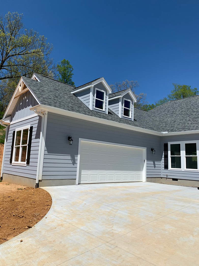 Two-story home with gray siding, white trim, attached garage, concrete driveway, and mature trees behind the house