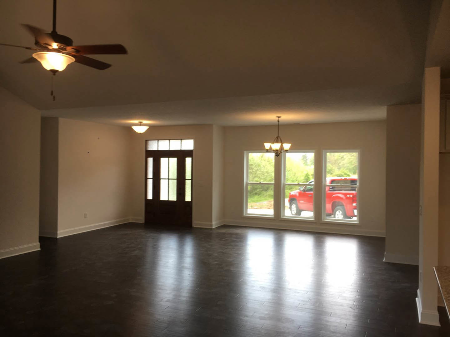 Living room with large window, red truck visible outside, light-colored walls, wood flooring, ceiling fan with light fixture