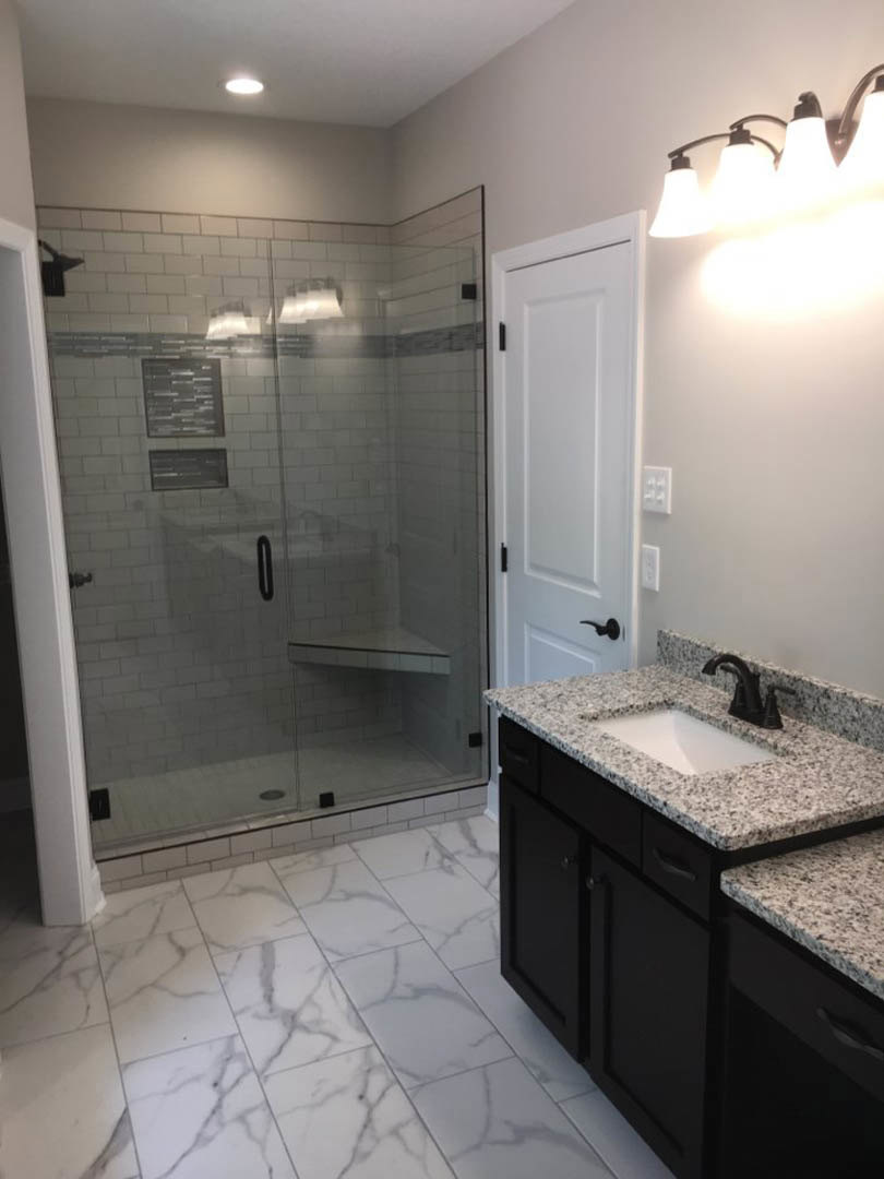 Bathroom with glass shower enclosure, built-in bench, white tile walls and floor with black trim, modern row light fixture above white sink and countertop