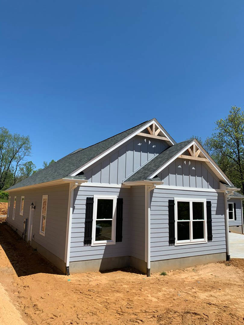 Partially built house with white siding, black shuttered window, dirt ground, and leafy trees under clear blue sky