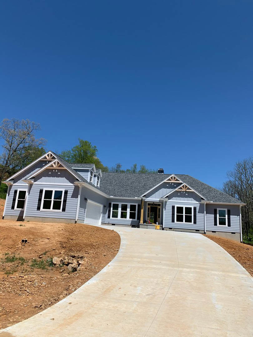 Two-story home with white siding, black shuttered windows, attached garage, concrete driveway bordered by dirt and rocks, small grassy patch, and covered porch under a cloudy sky.