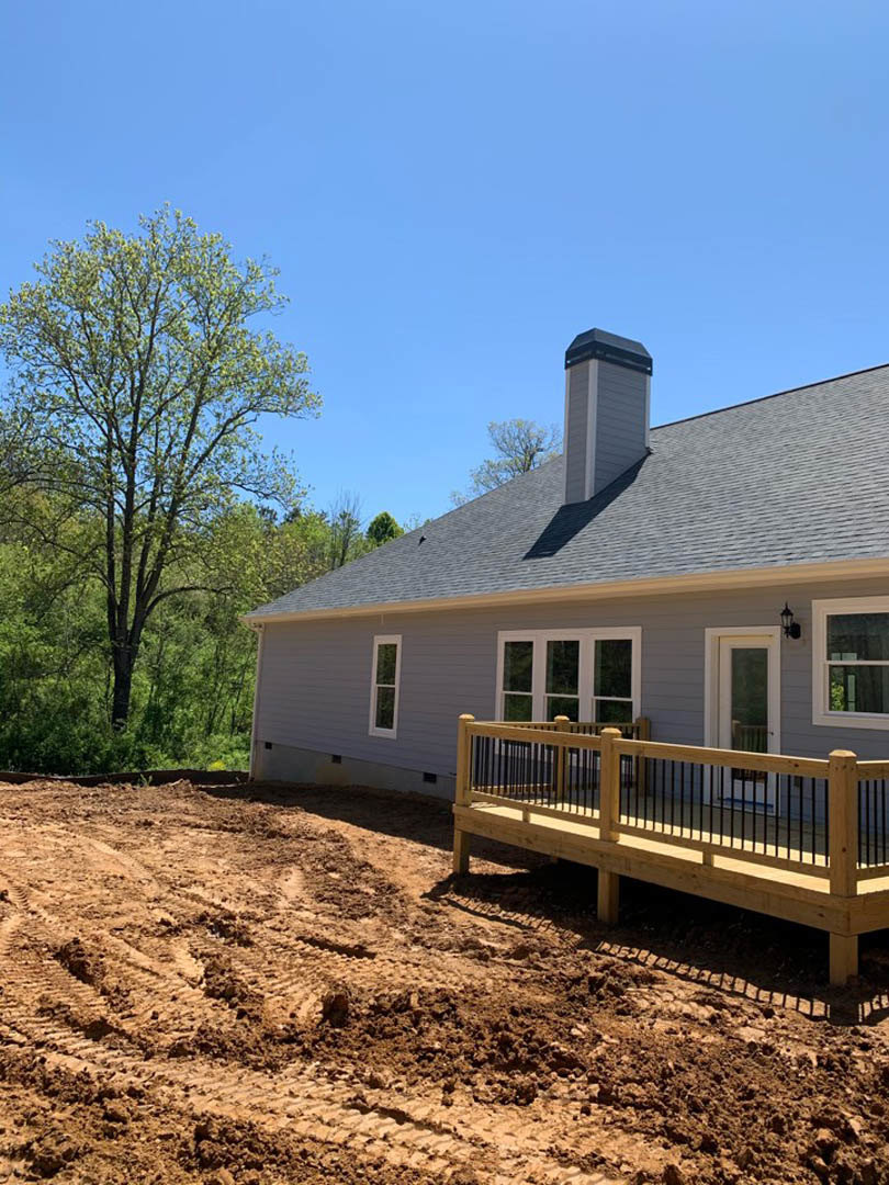 Two-story house with white-framed windows, wooden deck featuring black railing, dirt yard with tire tracks, and leafy green tree nearby