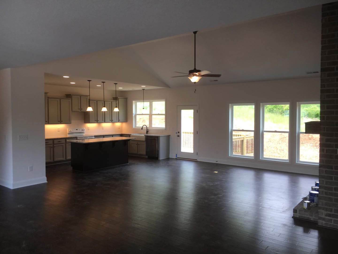 Spacious open-concept kitchen and living area featuring a dark wood floor, central kitchen island with black countertop, ceiling fan with light fixture, white door illuminated by