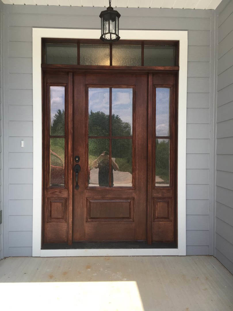 Wooden entry door with glass panes, metal handle, and outdoor reflection; adjacent window showing trees; interior lamp and hardwood floor visible