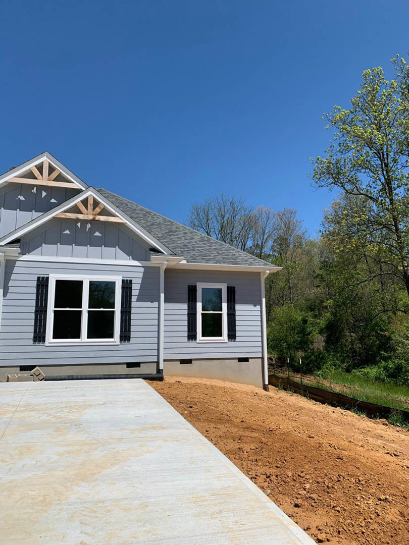 Two-story house under construction with exposed framing, concrete driveway, white window frames, surrounding trees, and dirt landscaping under a clear blue sky