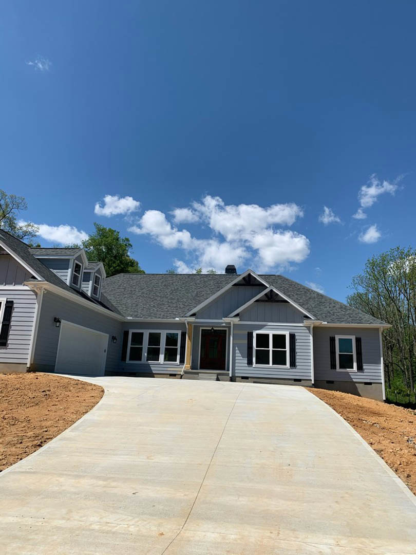 Concrete driveway leading to a two-story house with white-framed windows, gabled roof, and mature trees under a blue sky with scattered clouds