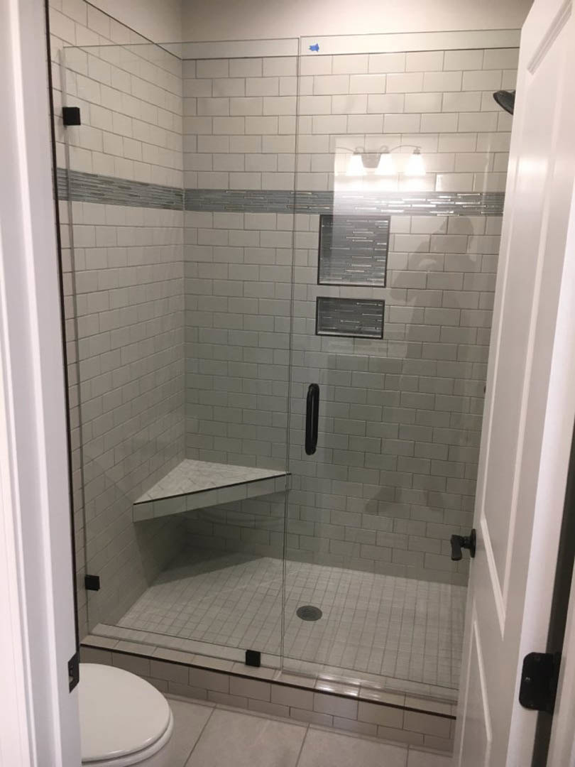 Frameless glass shower enclosure with built-in tiled shelf, adjacent white toilet seat, and partial view of bathroom window