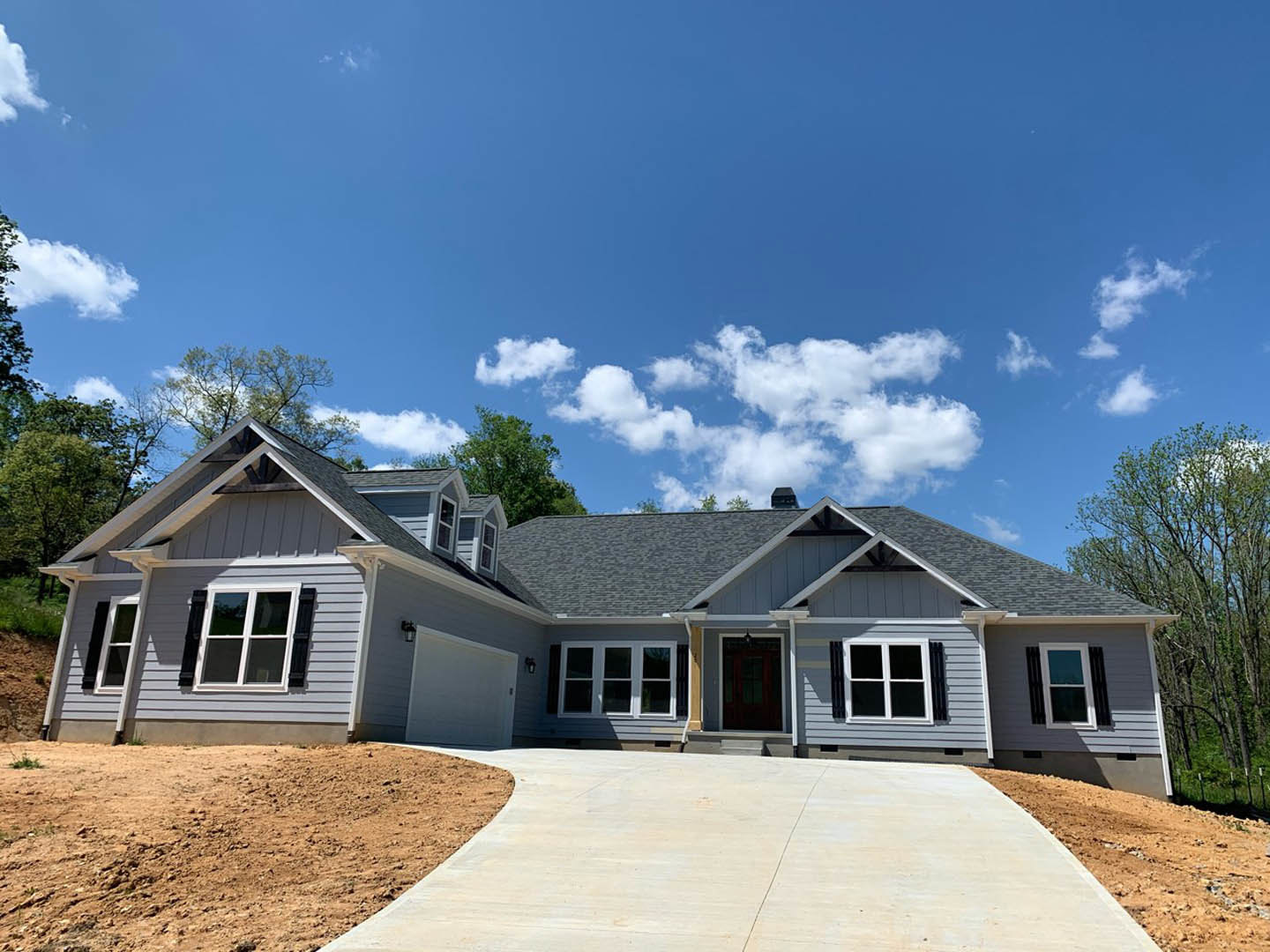 Two-story home with gray siding, white trim, large windows, concrete driveway, covered front porch, mature trees in the background, and blue sky with scattered clouds