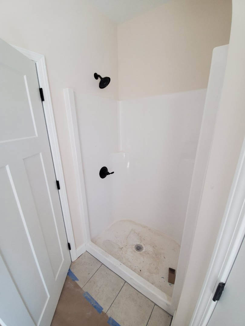Modern bathroom shower with black round shower head, white door partially visible, tile floor marked with blue tape, and spiral black drain cover.