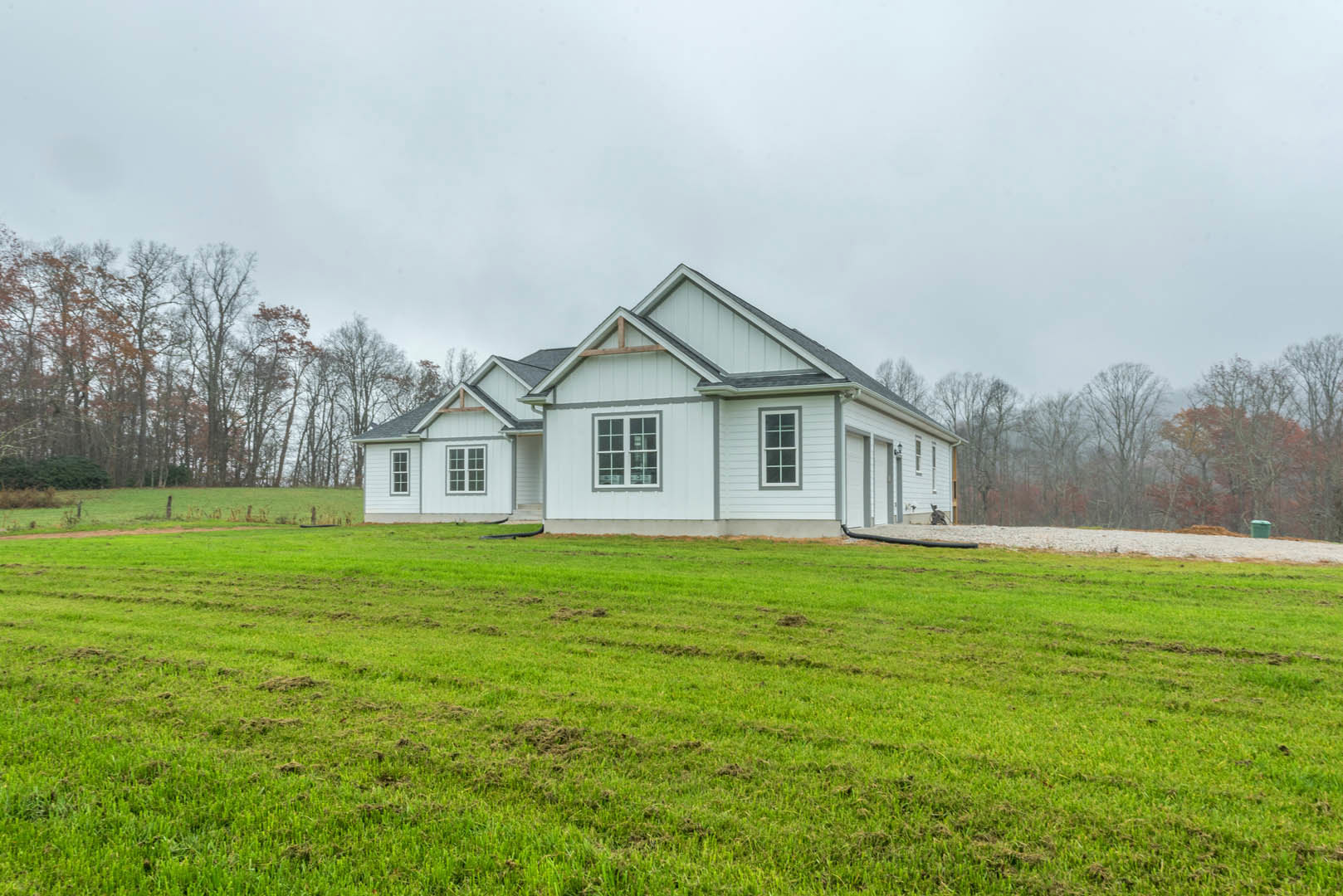 White farmhouse with gray trim and multiple-paned windows, set behind a large green lawn, surrounded by trees under a partly cloudy sky