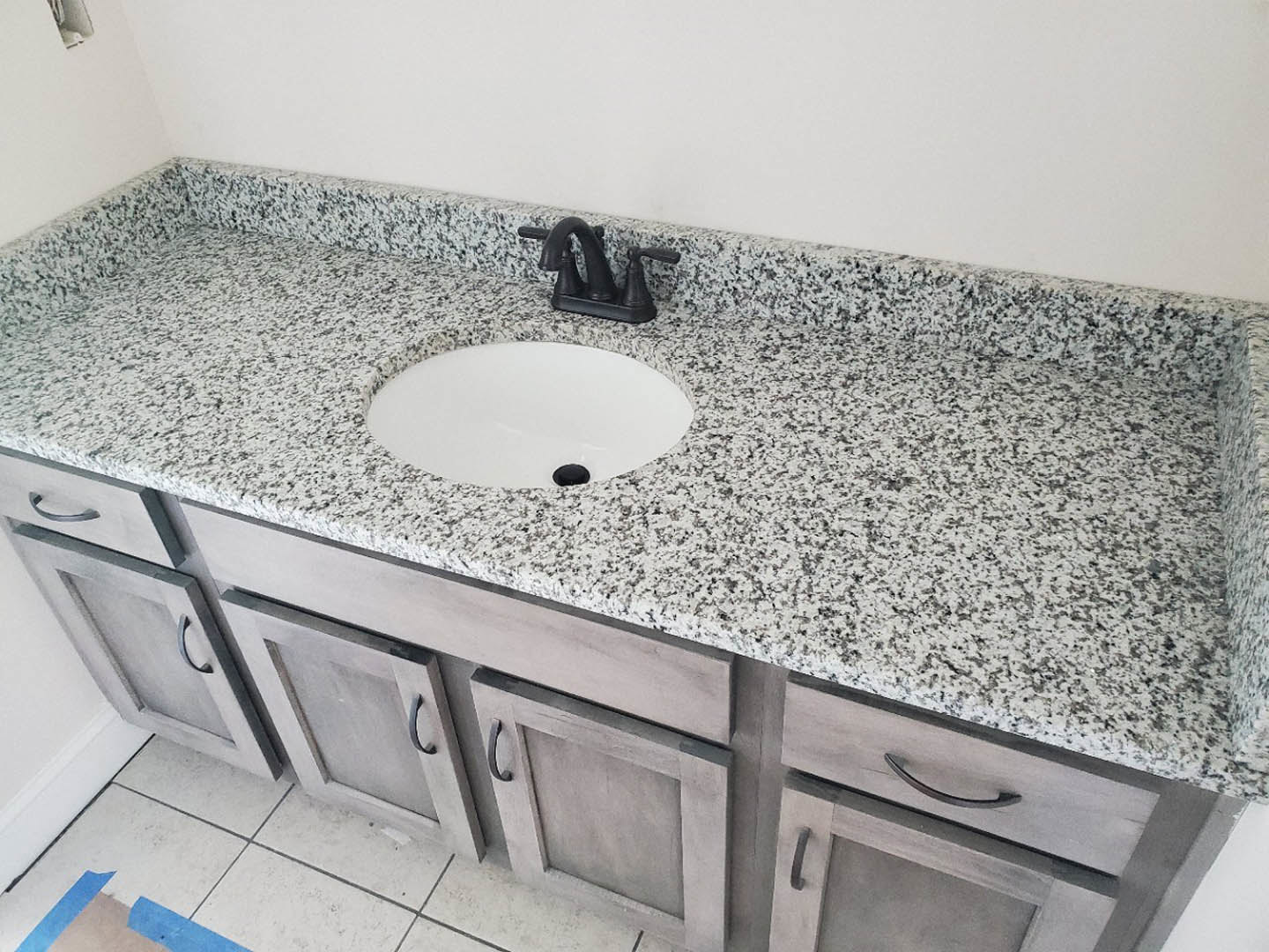 White rectangular sink with black faucet set on marbled countertop, dark cabinetry below, light tile backsplash and wall visible in modern bathroom.
