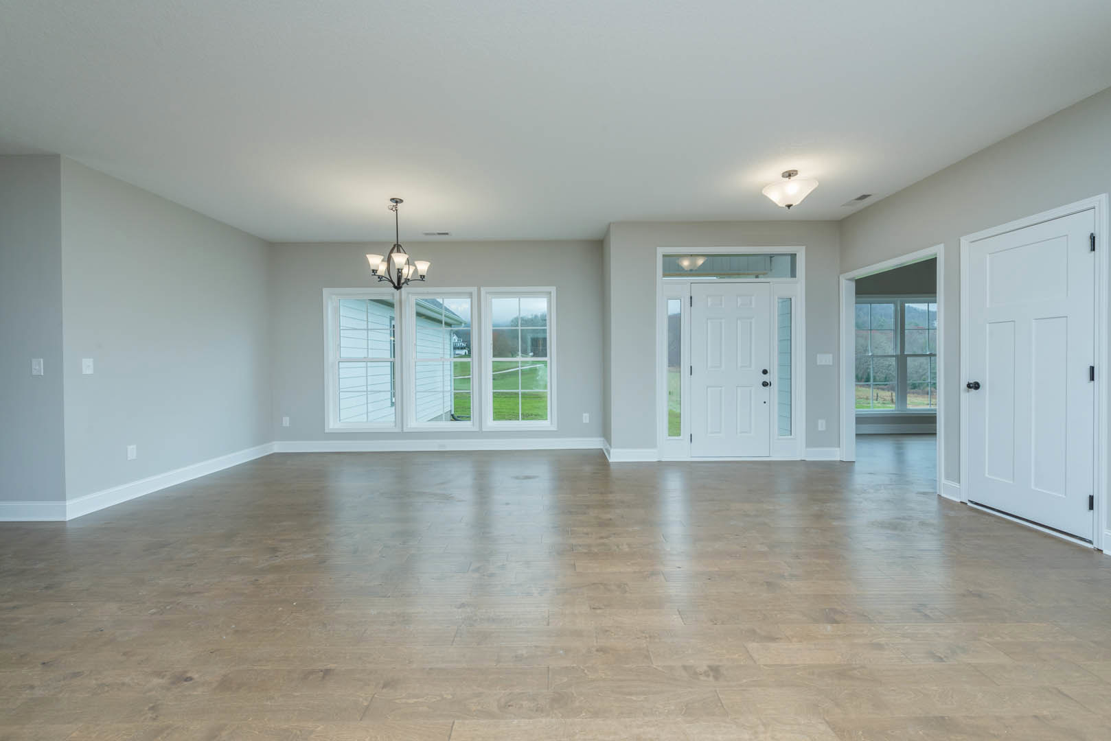 Hardwood floor room with white glass-paneled door featuring black knobs, large windows overlooking grassy field, white walls and ceiling