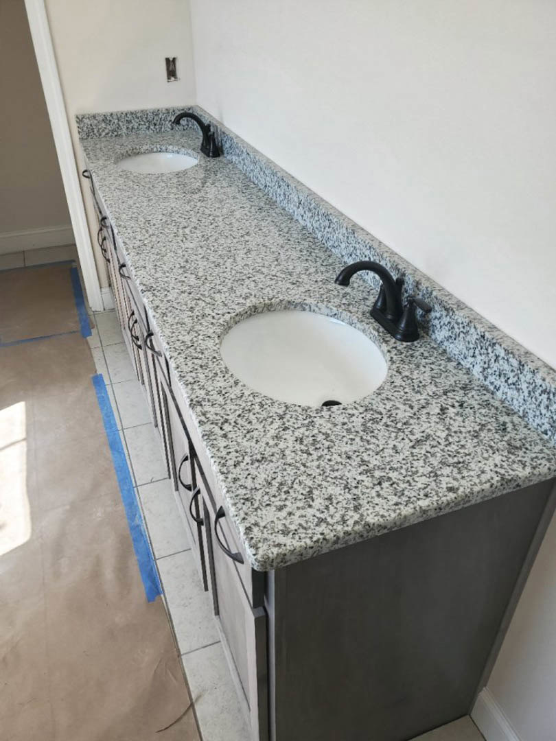 White bathroom sink set in a gray marble countertop with a matte black faucet, blue painter’s tape on the light tile floor, and neutral wall tiles in the background.