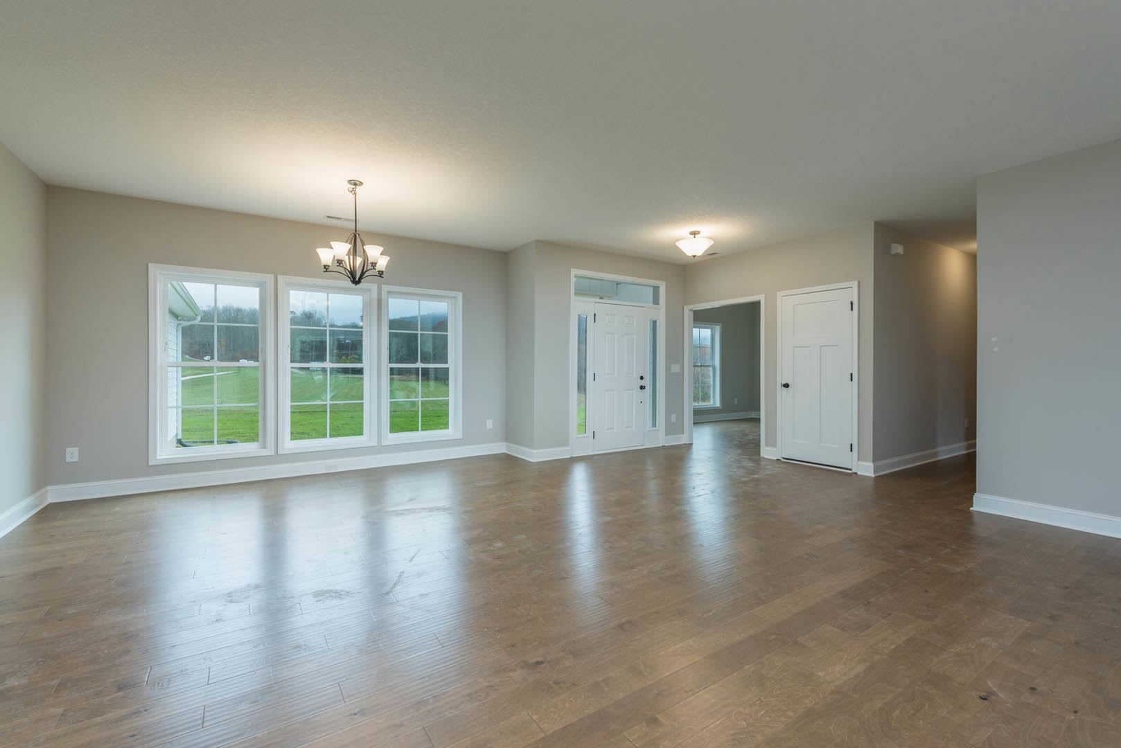Spacious room with hardwood floors, multiple large windows overlooking a green lawn, white walls, black door knobs, and a modern chandelier hanging from the ceiling