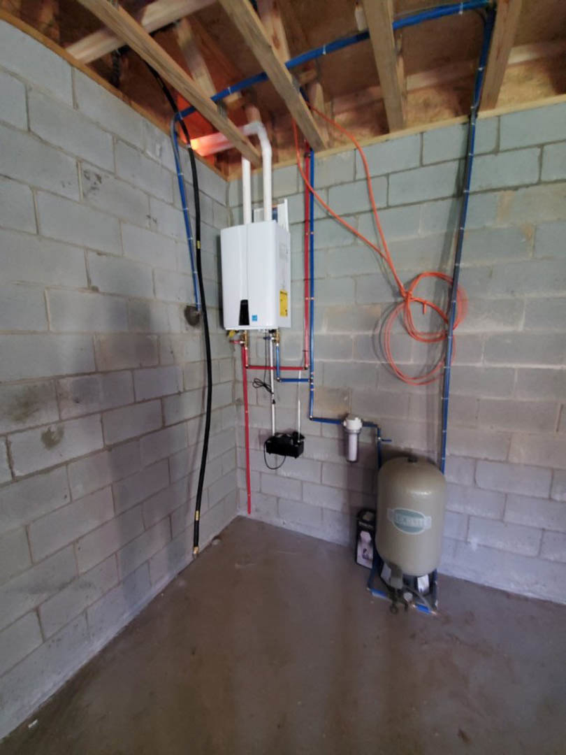White water heater with blue label installed in unfinished basement, surrounded by exposed wooden beams, pipes, concrete floor, and insulated walls.