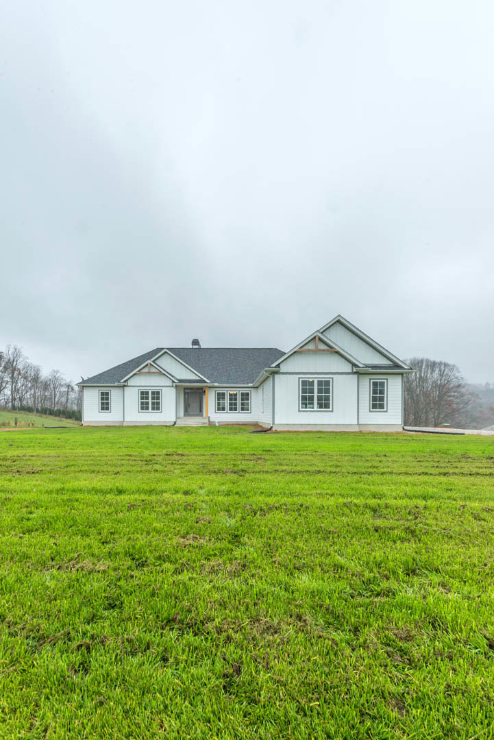 White farmhouse with gray roof set behind expansive green lawn, bordered by mature trees under partly cloudy sky