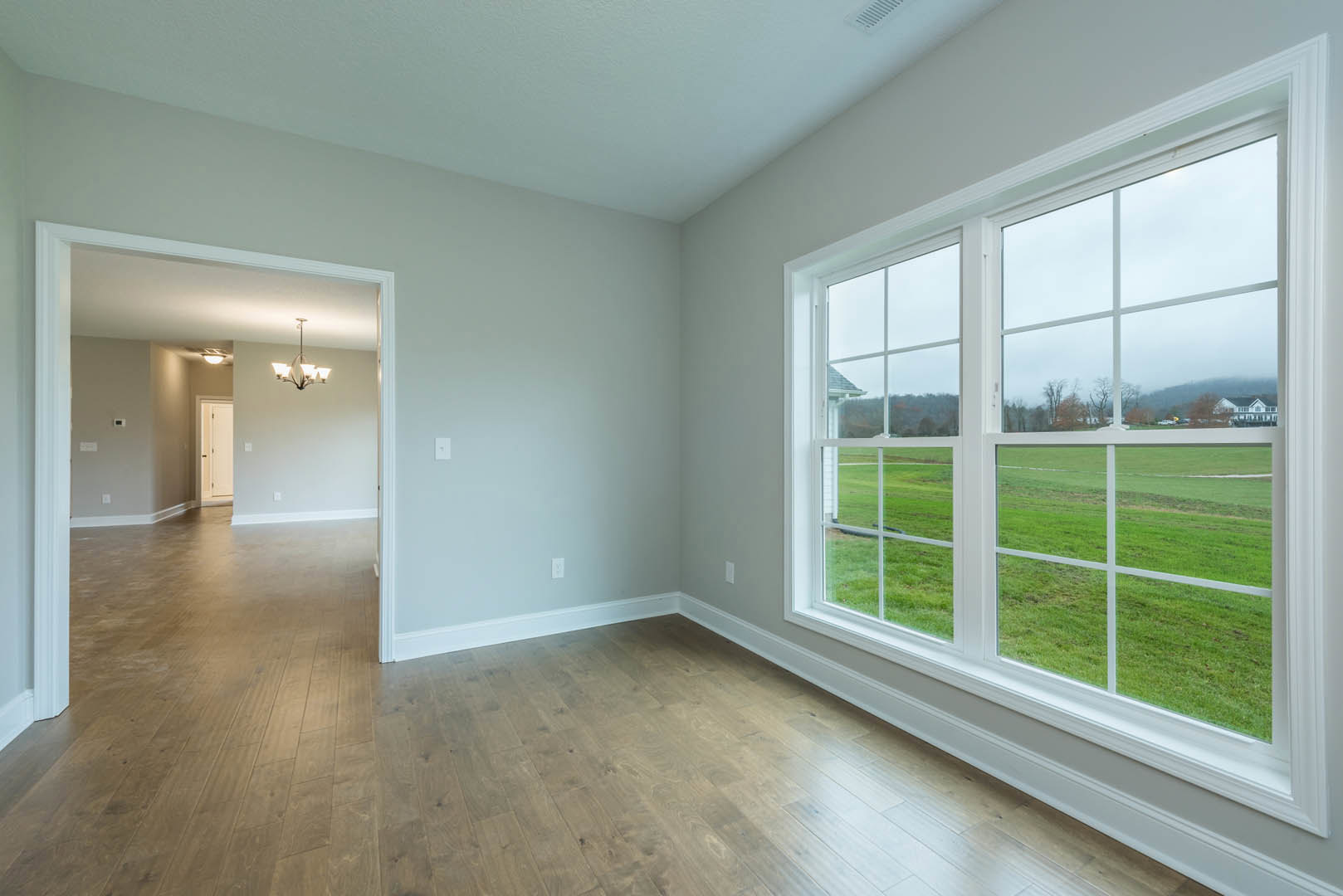 Spacious room featuring a large window overlooking a green grass field, wooden laminate flooring, neutral walls, and a modern chandelier fixture