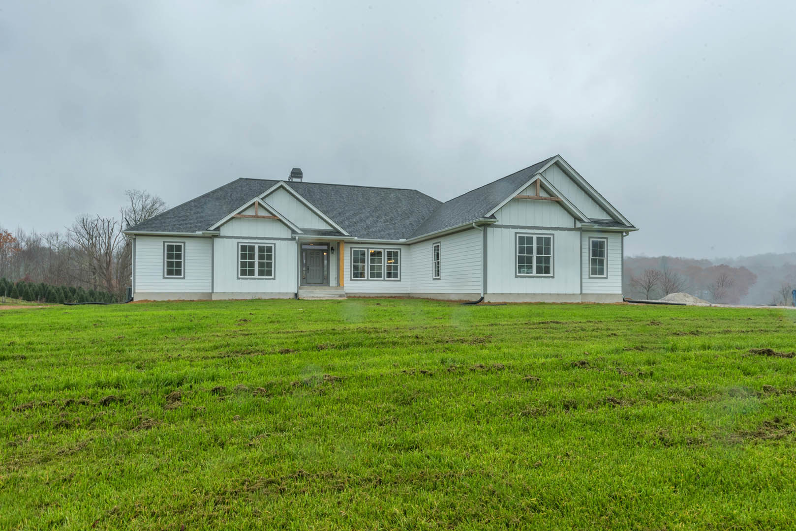 White farmhouse with grey roof and door, expansive green lawn in foreground, Southfork Ranch visible in background, cloudy sky overhead.