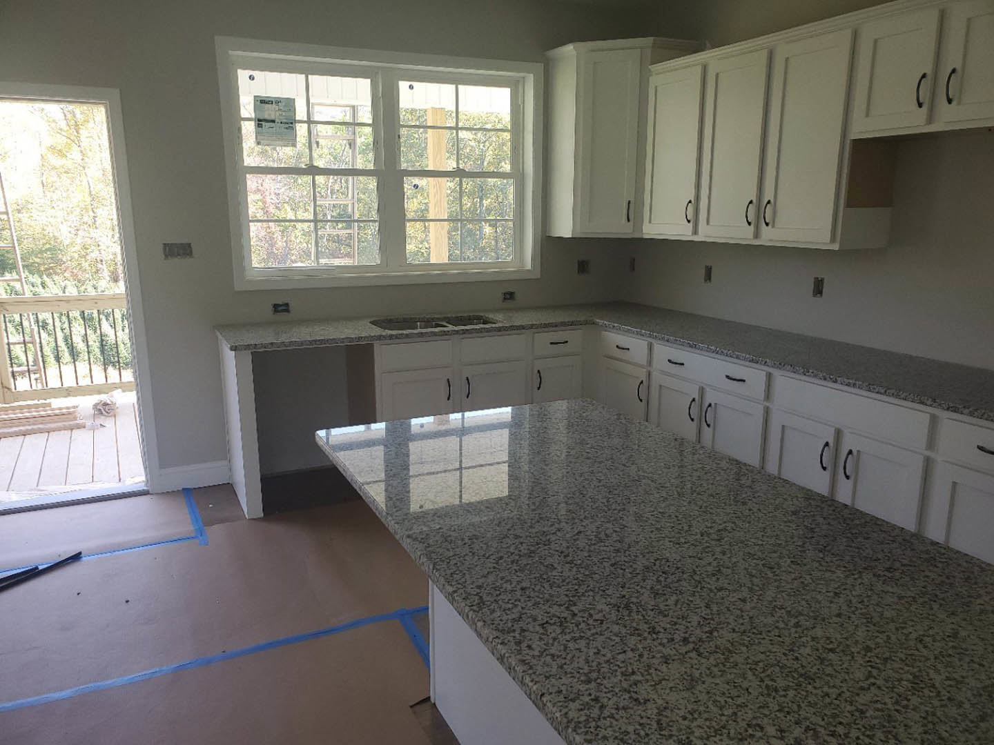 Granite kitchen countertops with white cabinets, stainless sink, tile flooring, large window showing trees, blue tape on floor, white sign attached to metal bar