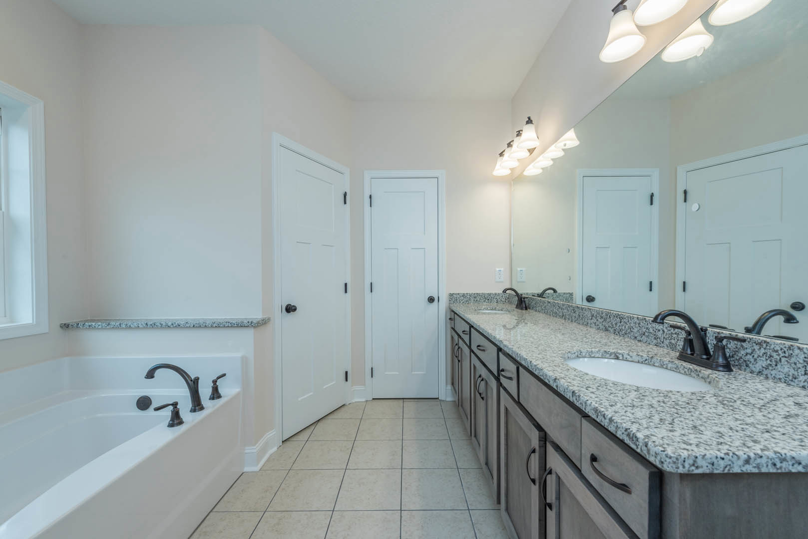 Bathroom with double sinks set in a white countertop, large frameless mirror above, white cabinetry with black hardware, white tile flooring, and a freestanding bathtub with chrome