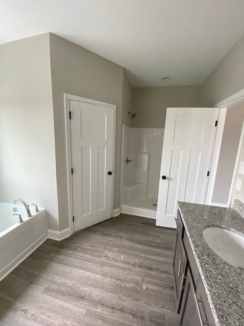 Bathroom with freestanding white bathtub, rectangular sink on wood vanity, black hardware, wood flooring, white door, light tile walls