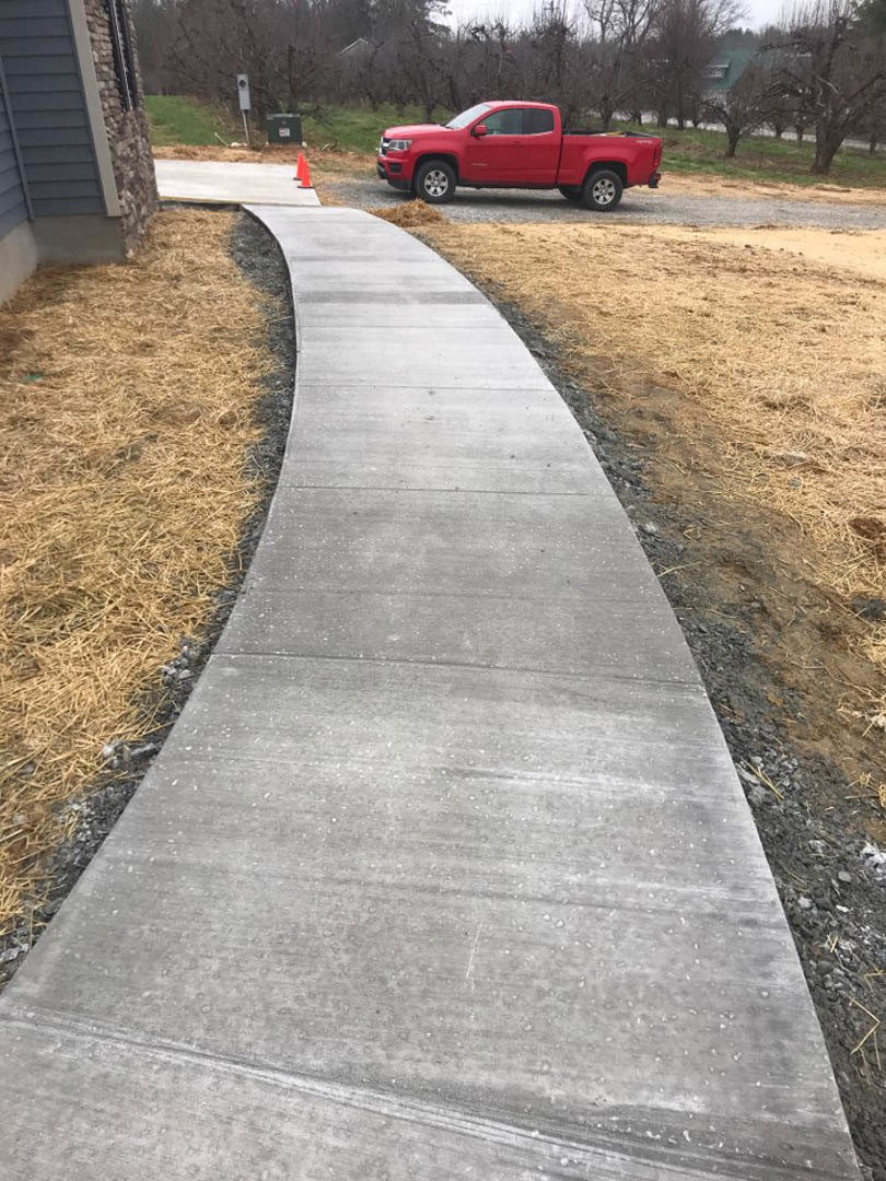 Concrete walkway bordered by green and dry grass, red truck parked on asphalt road, tree in background, close-up of house exterior