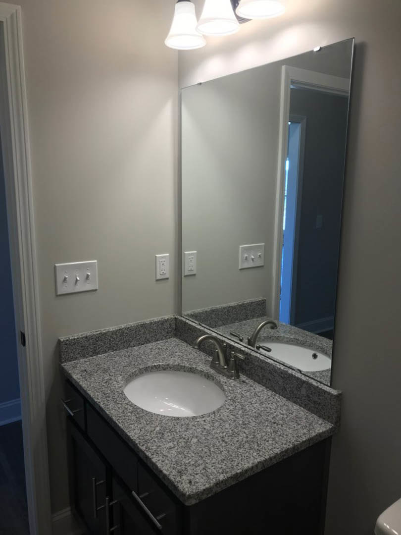 White bathroom with black speckled countertop, rectangular mirror above sink, three-switch white light panel, and cluster of white overhead lights