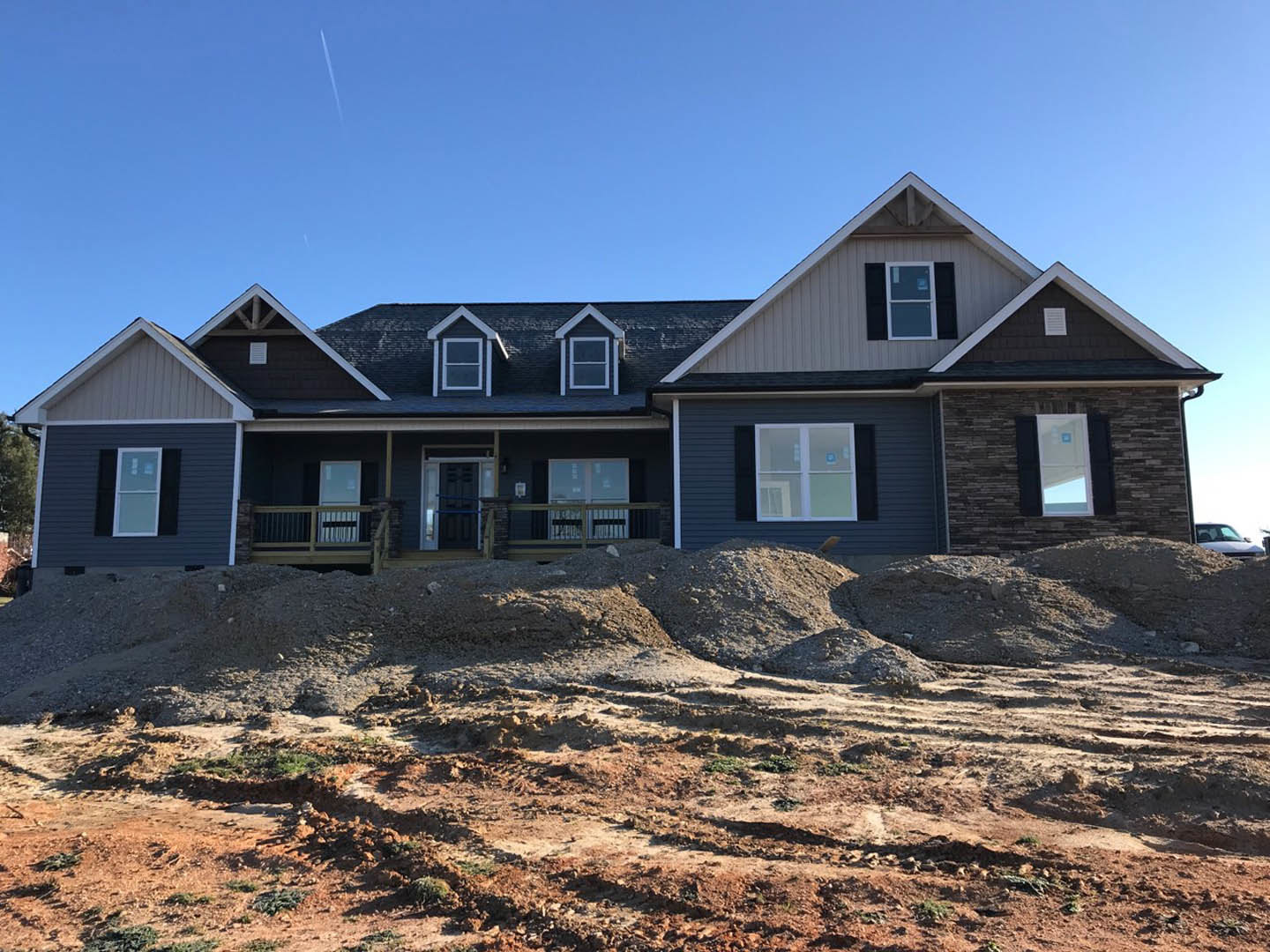 Two-story house under construction with blue roof, white-framed windows, metal railing, exposed siding, and piles of dirt covering the ground.