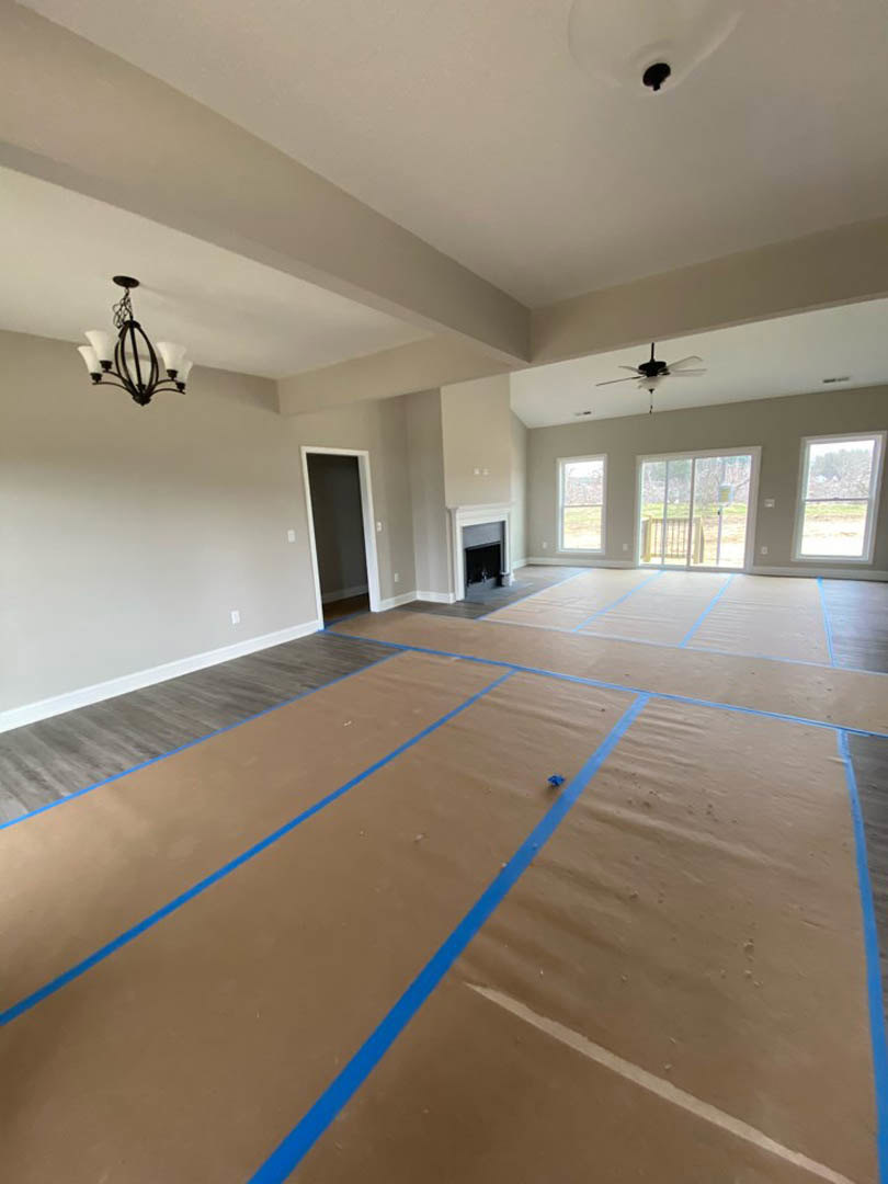 Room with blue tape marking layout on hardwood floor, black door with white trim, chandelier and ceiling fan overhead, windows showing fenced yard outside