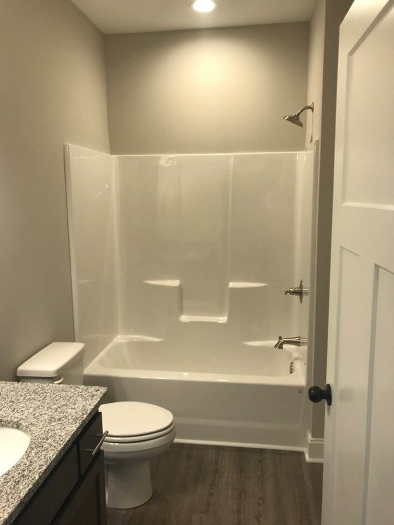 White bathtub with chrome faucet next to a toilet, light-colored tile walls, black door knob on white door, close-up of shower head and bathroom accessories