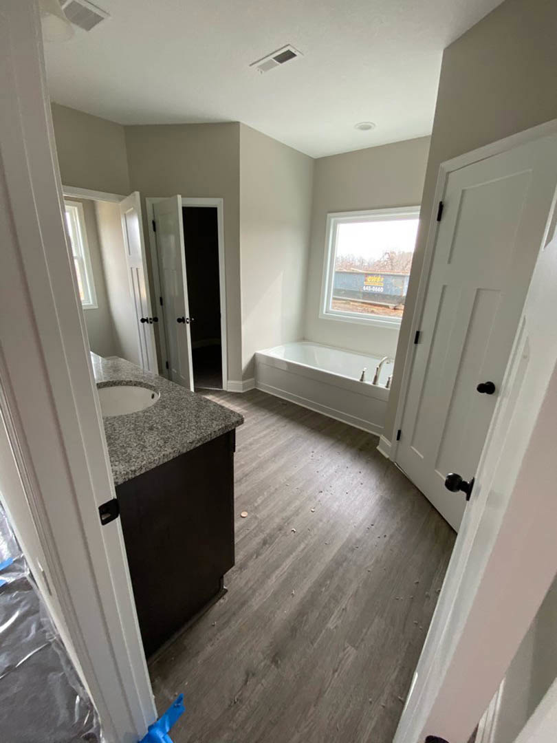 Bathroom with freestanding bathtub, white rectangular sink on wood vanity, wood laminate flooring, white door with black knob, large window overlooking outdoor container.