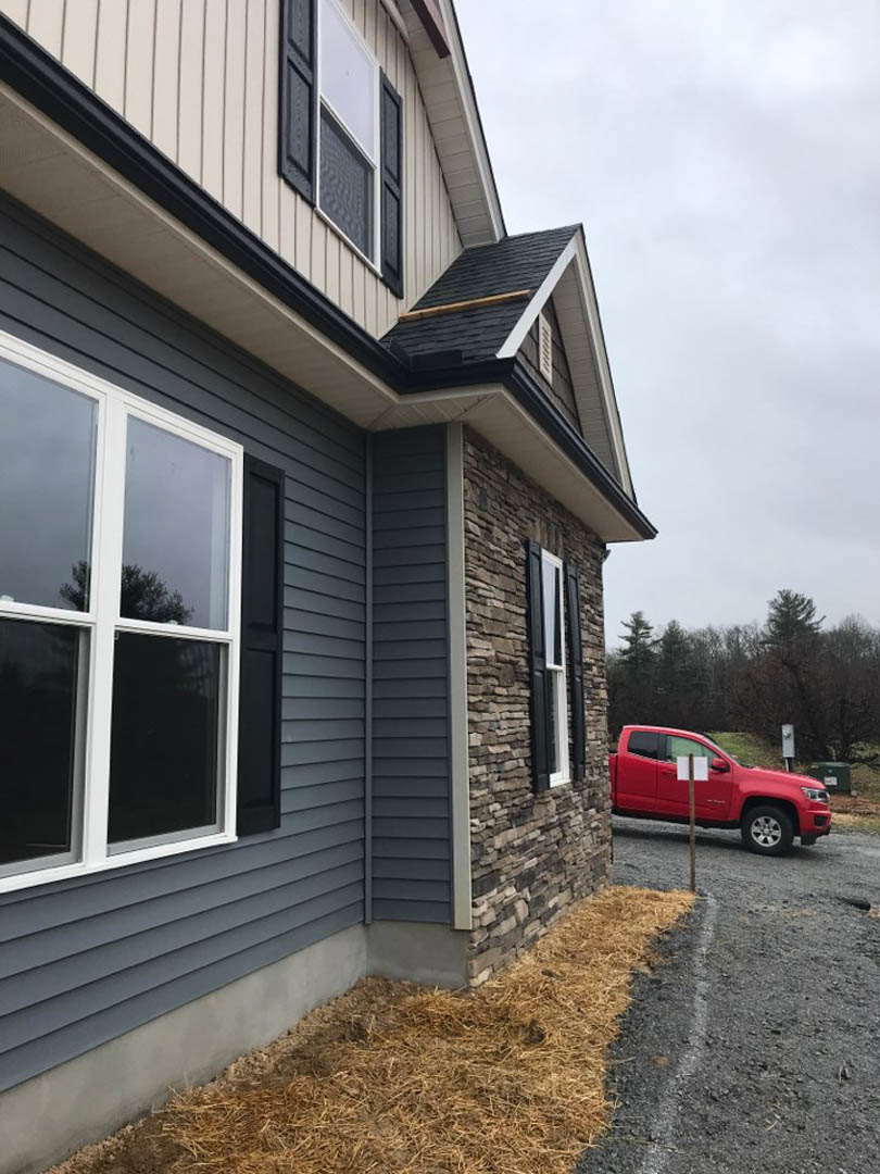 Two-story custom home with light siding, large windows, and gabled roof; red pickup truck parked on driveway in front; manicured lawn and mature trees visible in background