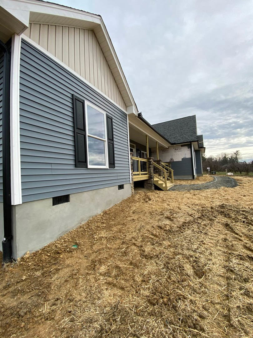 Two-story home with light siding, fenced grassy yard, dirt patch near porch, large windows, and cloudy sky overhead