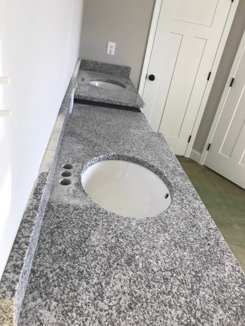 Granite bathroom countertop with two undermount sinks, chrome faucets, white cabinetry, wall-mounted mirror, and white door with black knobs