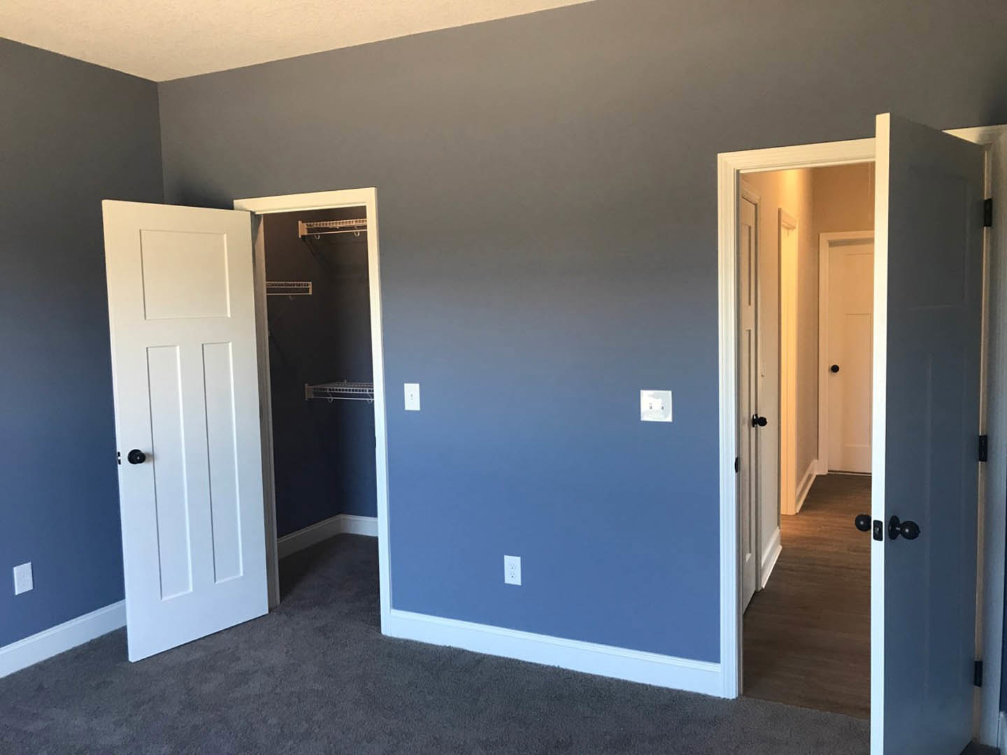 Bedroom with light walls and hardwood floor, two white doors open; one leads to a closet with a built-in shelf, both doors feature black knobs.