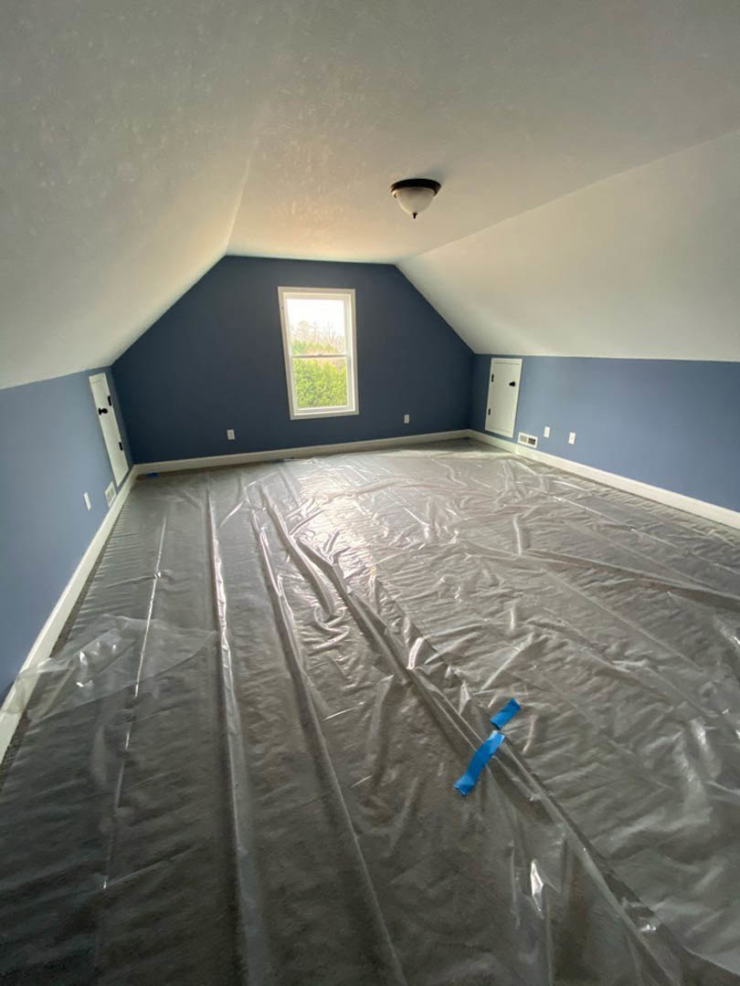 Room under renovation with plastic sheeting covering the floor, blue painter’s tape on a grey wall, large window showing trees outside, and ceiling light fixture visible