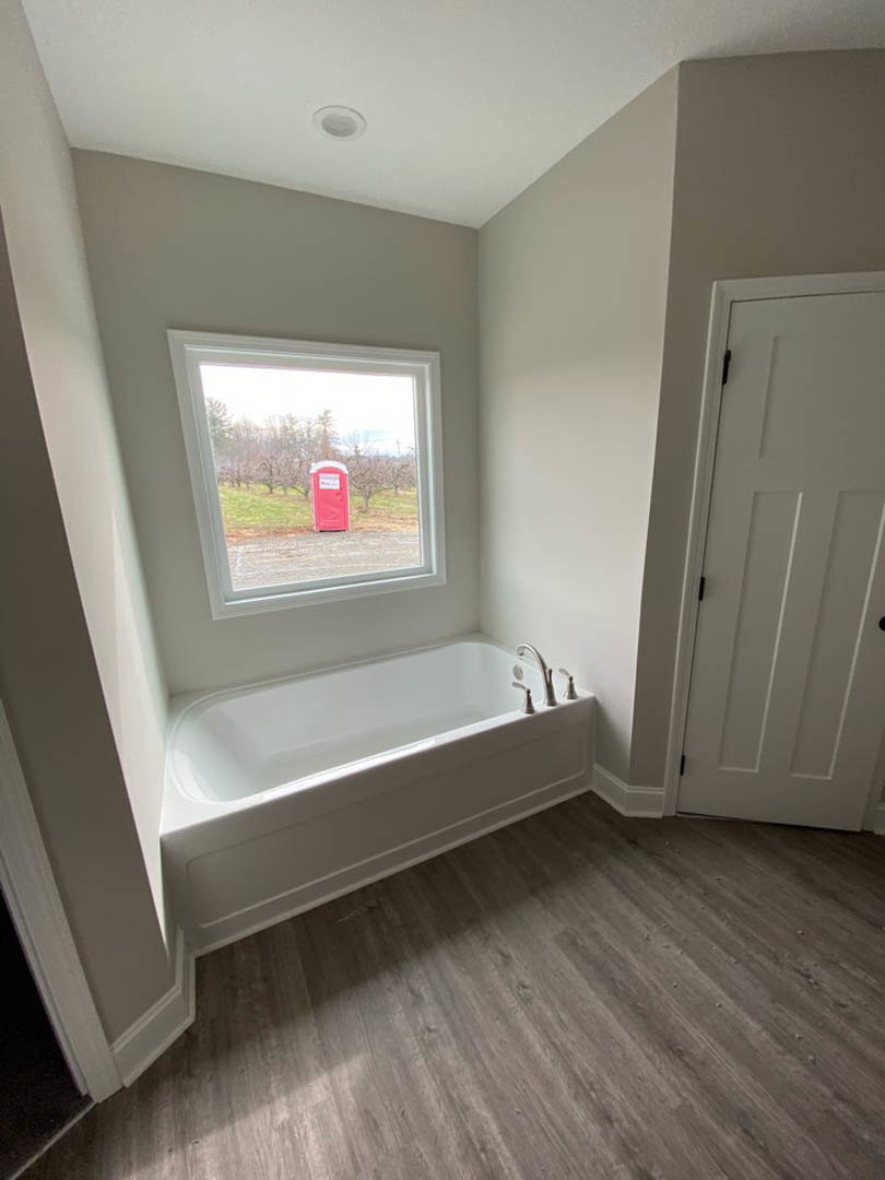Bathroom with freestanding white tub beneath window, wood flooring with white trim, white door with black handle, chrome faucet, and natural light illuminating neutral plaster