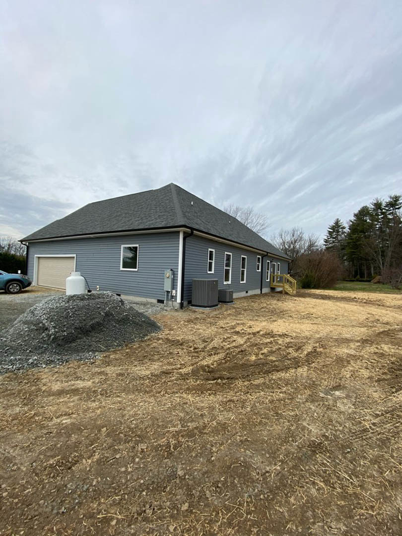 Grey-roofed house with a pile of dirt and gravel in the yard, white container with pipe near garage, man standing beside garage door, scattered rocks on bare ground, cloudy sky