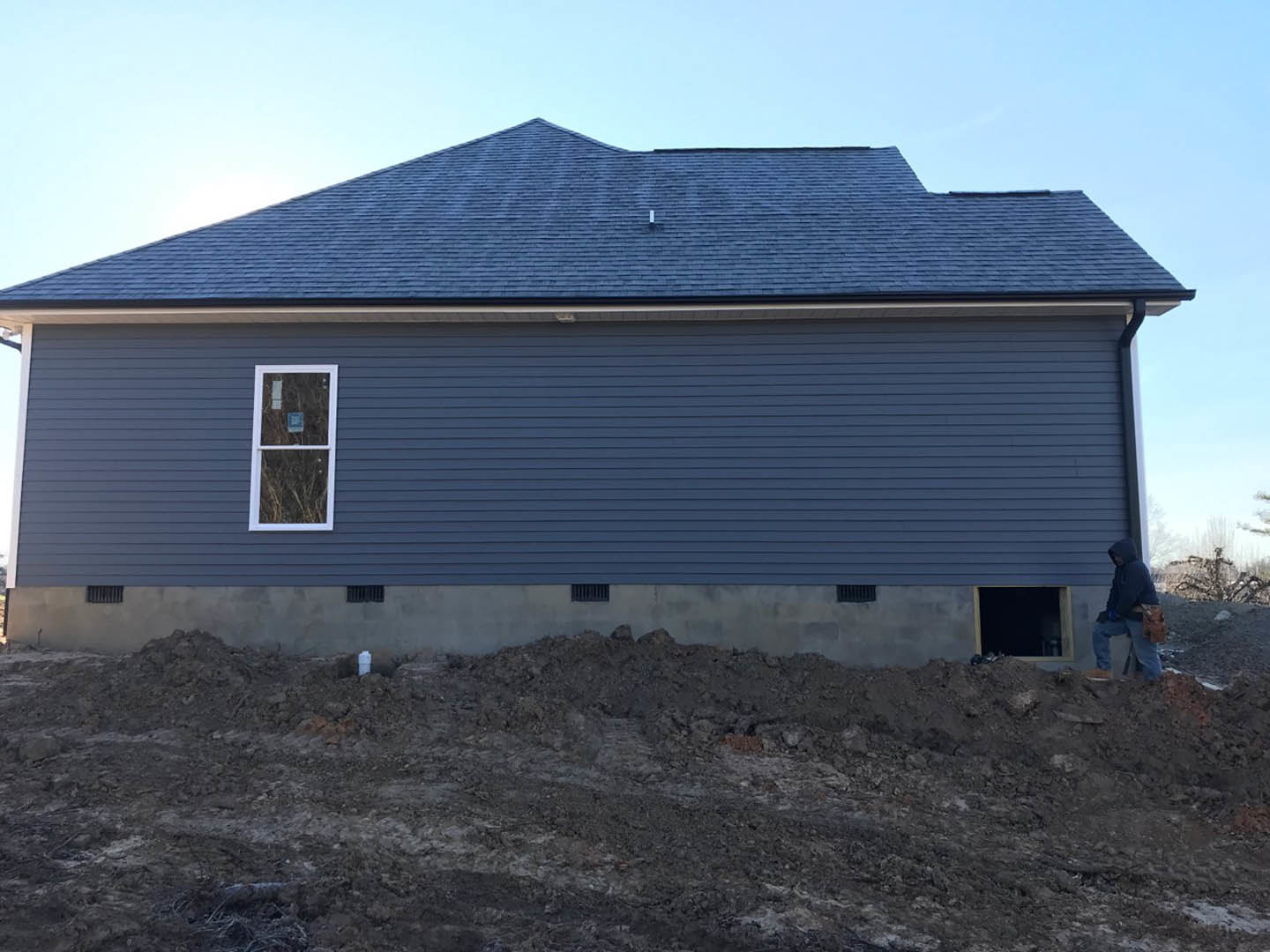 Partially finished house with exposed siding, large window displaying a construction sign, man in hoodie standing in front, pile of dirt against foundation wall, close-up view of