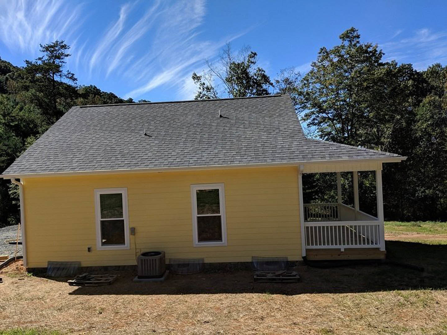 Yellow siding home with white framed windows, covered front porch featuring white railing, gray shingled roof, and small tree in the yard.